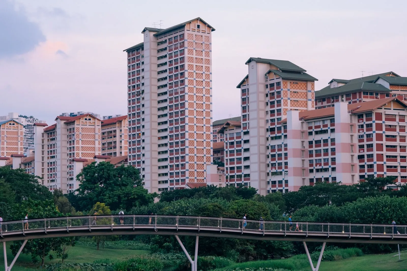 HDB blocks above Bishan-Ang Mo Kio Park in Singapore at dusk, with people crossing a footbridge below.