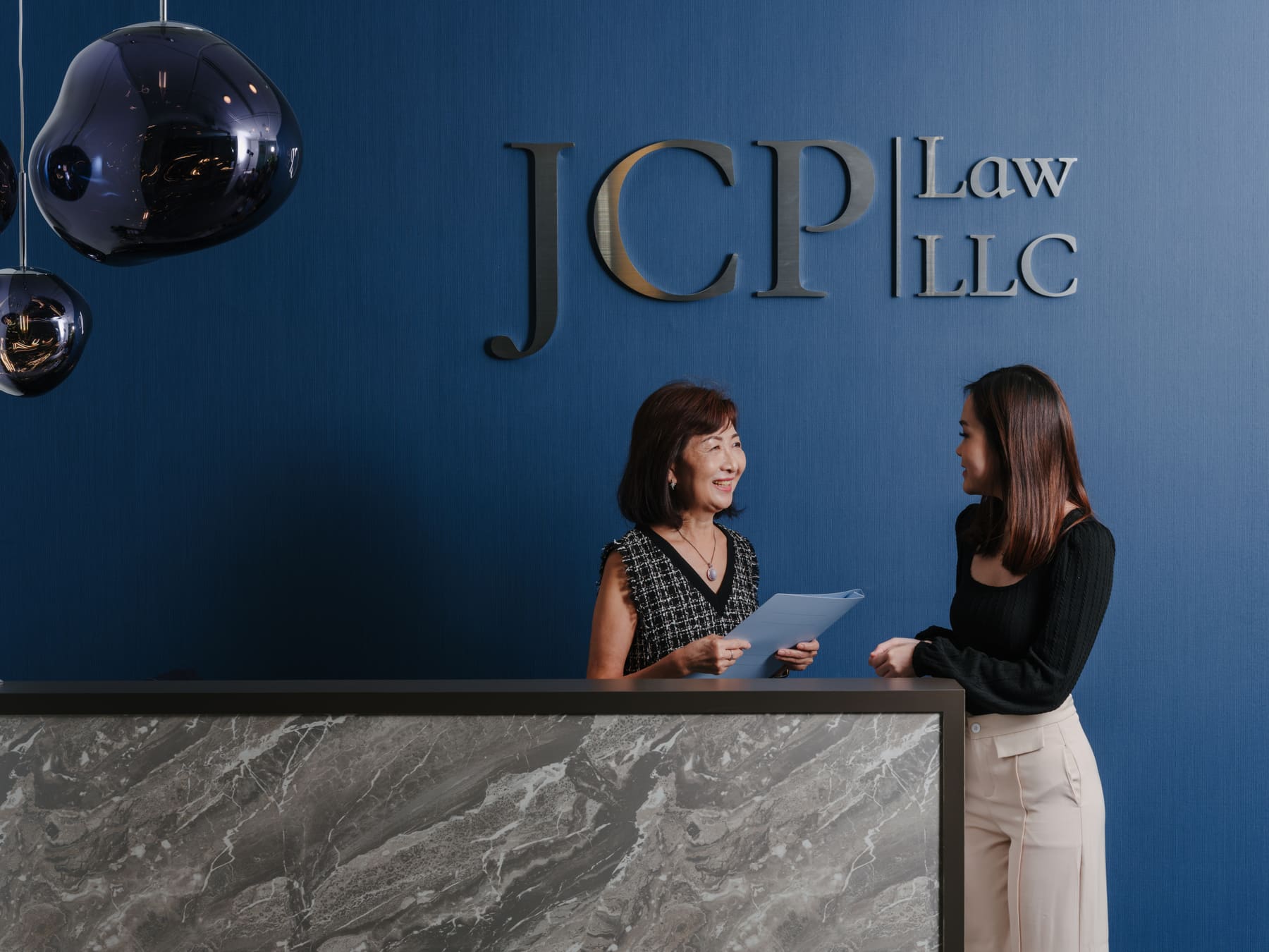 Two JCP Law staff members meeting at the reception desk, beneath the JCP Law LLC sign at 6 Battery Road, Singapore.